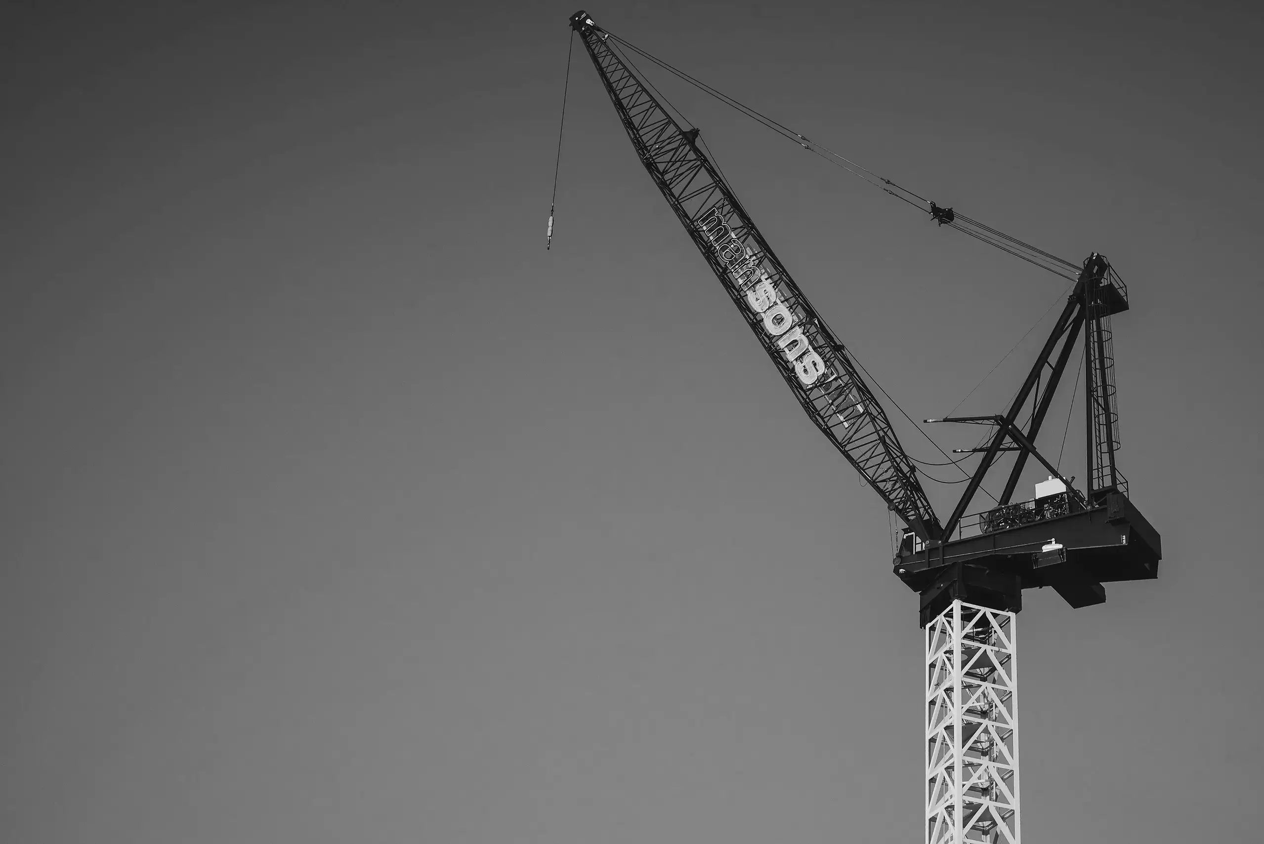 Schwarz-weiß-Fotografie eines großen Baukrans mit Gittermast und Ausleger vor klarem Himmel, seitlich mit dem Schriftzug „mansons“ am Ausleger – Symbol für Bauindustrie, Entwicklung und städtisches Wachstum.